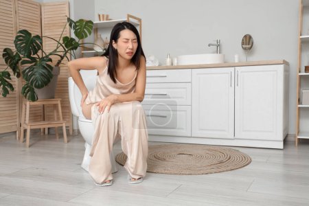 Joven mujer asiática con hemorroides sentado en el inodoro en el baño