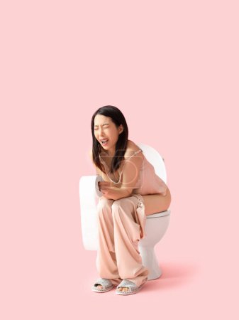 Photo for Young Asian woman in pajamas with hemorrhoids sitting on toilet bowl against pink background - Royalty Free Image