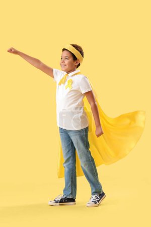 Téléchargez les photos : Mignon petit garçon afro-américain en costume de super-héros avec ruban jaune sur fond de couleur. Concept de sensibilisation au cancer chez l'enfant - en image libre de droit
