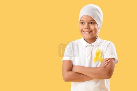 Téléchargez les photos : Mignon petit garçon afro-américain après la chimiothérapie avec ruban jaune sur fond de couleur. Concept de sensibilisation au cancer chez l'enfant - en image libre de droit
