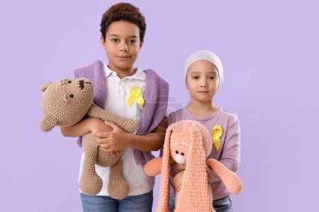 Téléchargez les photos : Petits enfants mignons avec des rubans jaunes et des jouets sur fond lilas. Concept de sensibilisation au cancer chez l'enfant - en image libre de droit