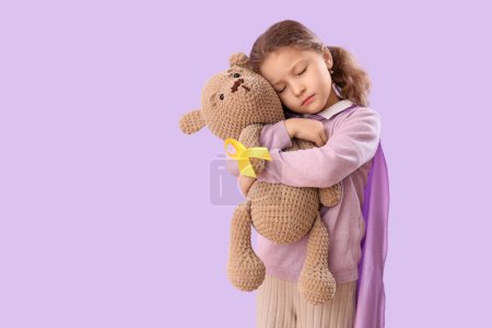 Téléchargez les photos : Jolie petite fille avec ruban jaune et ours en peluche sur fond lilas. Concept de sensibilisation au cancer chez l'enfant - en image libre de droit