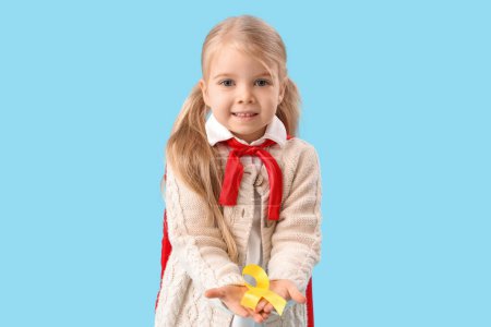Téléchargez les photos : Petite fille mignonne en costume de super héros avec ruban jaune sur fond bleu. Concept de sensibilisation au cancer chez l'enfant - en image libre de droit