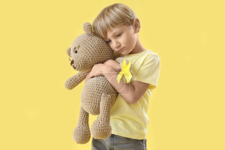 Téléchargez les photos : Mignon petit garçon avec ruban jaune et ours en peluche sur fond de couleur. Concept de sensibilisation au cancer chez l'enfant - en image libre de droit