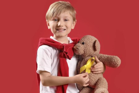 Téléchargez les photos : Joli petit garçon avec ruban jaune et ours en peluche sur fond rouge. Concept de sensibilisation au cancer chez l'enfant - en image libre de droit