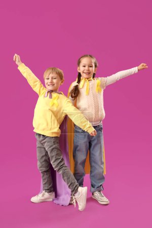 Téléchargez les photos : Petits enfants mignons en costume de super-héros avec des rubans jaunes sur fond violet. Concept de sensibilisation au cancer chez l'enfant - en image libre de droit