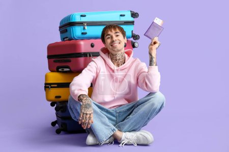 Foto de Joven feliz con pasaporte y billete sentado cerca de maletas contra fondo lila - Imagen libre de derechos