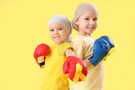 Téléchargez les photos : Jolies petites filles en gants de boxe avec des rubans jaunes sur fond de couleur. Concept de sensibilisation au cancer chez l'enfant - en image libre de droit