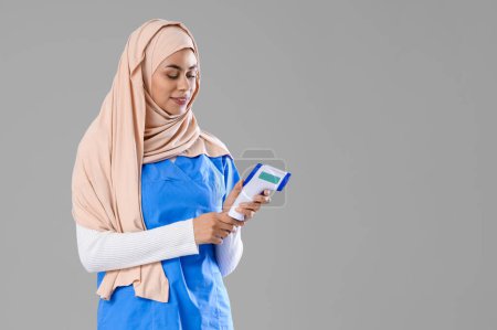 Female Muslim medical intern with infrared thermometer on light background