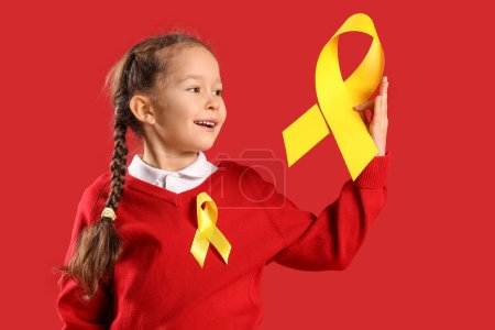 Téléchargez les photos : Jolie petite fille avec des rubans jaunes sur fond rouge. Concept de sensibilisation au cancer chez l'enfant - en image libre de droit