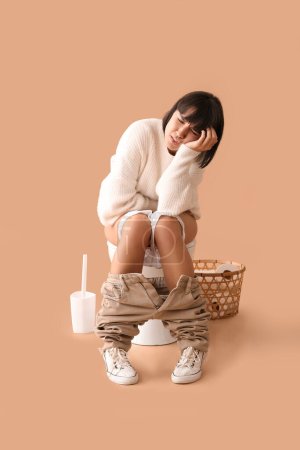 Foto de Mujer joven con dolor de estómago sentada en el inodoro sobre fondo beige - Imagen libre de derechos
