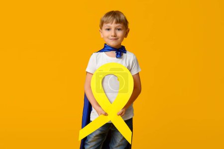 Téléchargez les photos : Mignon petit garçon en costume de super-héros avec ruban jaune sur fond de couleur. Journée internationale du cancer chez les enfants - en image libre de droit