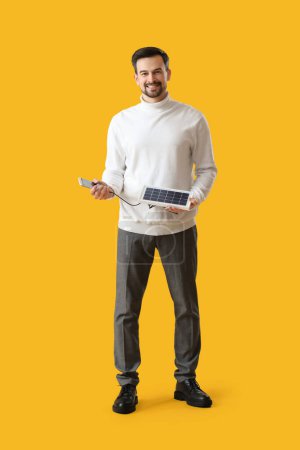 Foto de Hombre guapo con panel solar portátil cargando teléfono móvil sobre fondo amarillo - Imagen libre de derechos