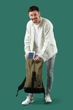 Foto de Hombre guapo poniendo el panel solar portátil en la mochila sobre fondo verde - Imagen libre de derechos