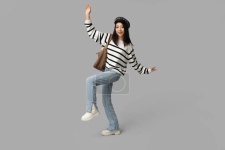 Schöne junge glückliche Asiatin in stylischen Jeans mit Accessoires auf grauem Hintergrund
