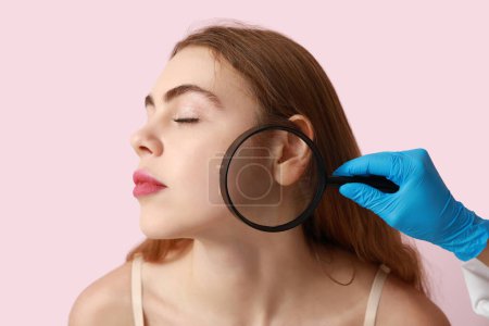Téléchargez les photos : Dermatologue examinant les taupes sur le visage d'une jeune femme avec loupe sur fond rose, gros plan - en image libre de droit