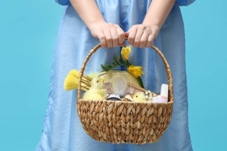 Beautiful young woman with basket full of decorative cosmetics on blue background, closeup
