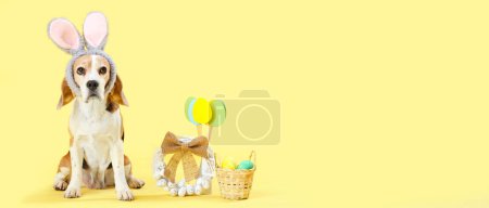 Cute Beagle dog with bunny ears, Easter eggs and wreath on yellow background with space for text