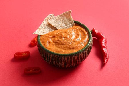 Bowl of tasty muhammara and chili peppers on red background