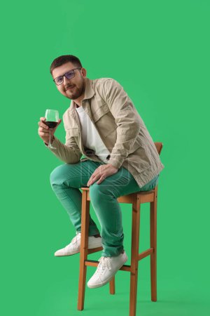 Photo for Young sommelier with glass of wine sitting on wooden chair against green background - Royalty Free Image
