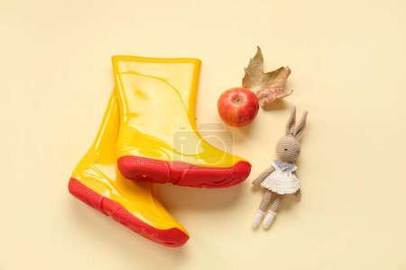 Foto de Botas de goma amarillas con conejito de juguete, manzana y hoja de otoño sobre fondo de color - Imagen libre de derechos