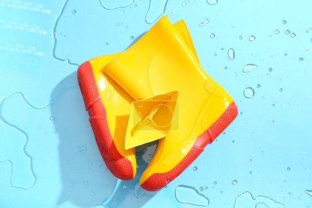 Foto de Botas de goma amarillas y barco de papel en agua sobre fondo azul - Imagen libre de derechos