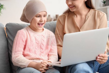 Téléchargez les photos : Petite fille après la chimiothérapie avec sa mère utilisant un ordinateur portable à la maison. Journée internationale du cancer chez les enfants - en image libre de droit