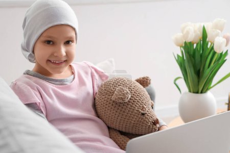 Téléchargez les photos : Petite fille après chimiothérapie avec ours jouet en utilisant un ordinateur portable à la maison, gros plan. Journée internationale du cancer chez les enfants - en image libre de droit