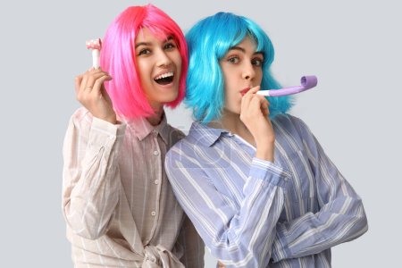 Beautiful young happy women in wigs with party whistles on grey background. April Fools Day celebration