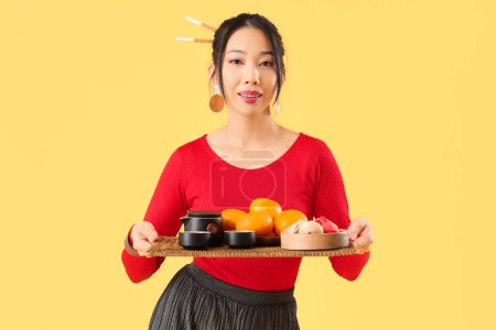 Photo for Young Asian woman holding tray with tea, mandarins and fortune cookies on yellow background. Chinese New Year celebration - Royalty Free Image