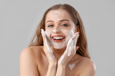 Beautiful young woman washing her face with foam on light background, closeup