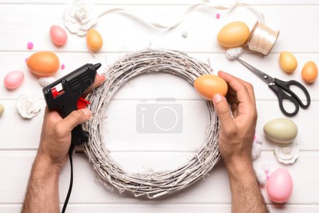 Téléchargez les photos : Homme créant une couronne de Pâques avec des œufs sur une table en bois blanc - en image libre de droit