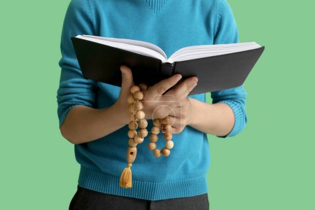 Little Muslim boy with Quran and praying beads on green background, closeup