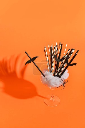 Glass with drinking straws and decor for Halloween celebration on orange background