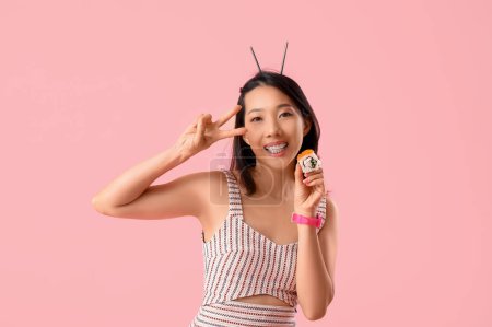 Pretty young Asian woman with tasty Philadelphia roll showing victory gesture on pink background