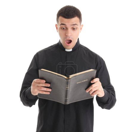 Shocked young priest reading Holy Bible on white background