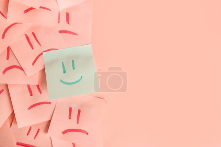Ein Blatt Papier mit gezeichnetem glücklichen Gesicht inmitten von Blättern mit traurigen auf rosa Hintergrund. Konzept der Einzigartigkeit