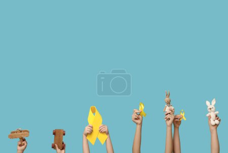 Téléchargez les photos : Les mains des enfants tenant des jouets et des rubans jaunes sur fond bleu. Journée internationale du cancer chez les enfants - en image libre de droit