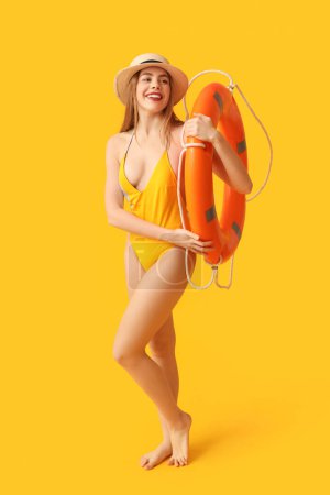 Pretty young woman in tank swimsuit and with lifebuoy on yellow background