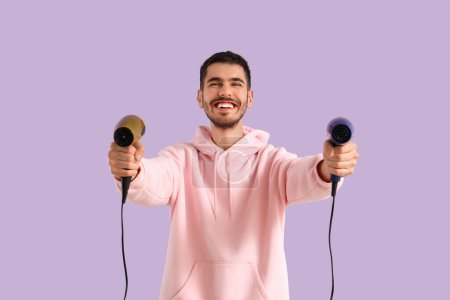 Handsome young man with hair dryers on lilac background
