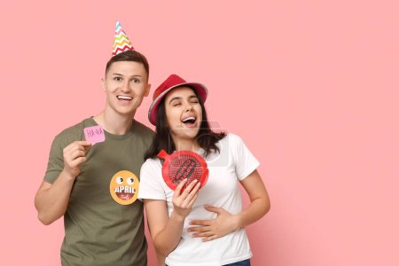 Young friends in funny disguise with paper stickers and whoopee cushion on pink background. April fool's day celebration