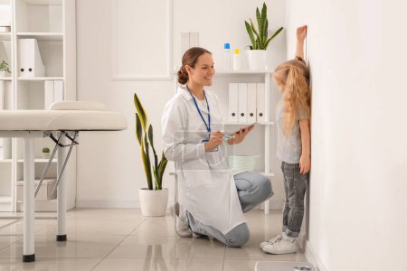 Kinderarzt misst Körpergröße von süßem kleinen Mädchen nahe weißer Wand in Klinik