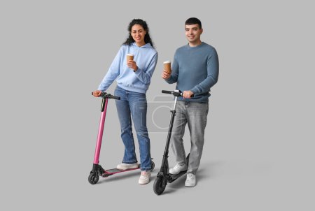 Foto de Hermosa pareja joven con modernos scooters de patada eléctrica y tazas de papel de café sobre fondo gris - Imagen libre de derechos