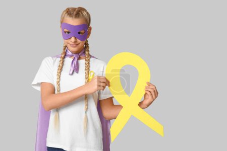 Téléchargez les photos : Adolescente mignonne en costume de super-héros avec ruban jaune sur fond gris. Journée internationale du cancer chez les enfants - en image libre de droit