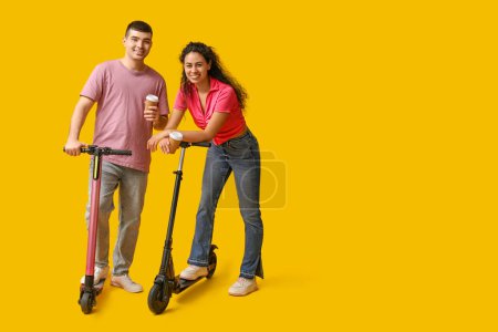 Téléchargez les photos : Beau jeune couple avec des scooters électriques modernes et des tasses en papier de café sur fond jaune - en image libre de droit