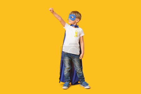 Téléchargez les photos : Mignon petit garçon en costume de super-héros avec ruban jaune sur fond de couleur. Journée internationale du cancer chez les enfants - en image libre de droit