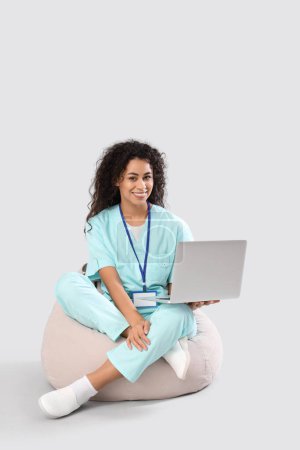 Female African-American medical intern with laptop sitting on light background