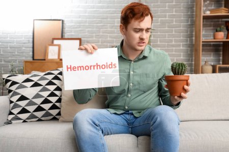 Young man holding paper with word HEMORRHOIDS and cactus on sofa at home