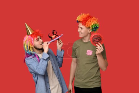 Young friends in funny disguise with whistle and whoopee cushion on red background. April fool's day celebration