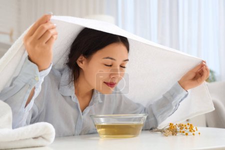 Young Asian woman with towel doing steam inhalation at table in bedroom